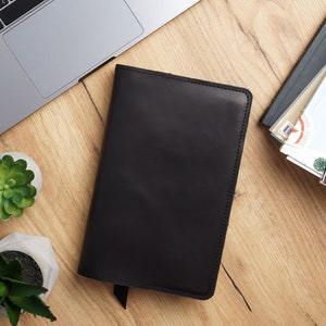 May include: A black leather notebook with a stitched edge and a flap closure. The notebook is lying on a wooden surface.