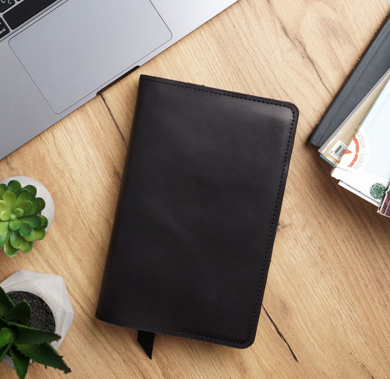 May include: A black leather notebook with a stitched edge and a flap closure. The notebook is lying on a wooden surface.