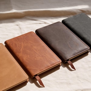 Pode incluir: Três cadernos retangulares com capas lisas em castanho claro, castanho médio e cinzento-carvão escuro. Cada caderno apresenta bordas costuradas e um marcador de fita de tecido a condizer que se estende da borda inferior da capa.