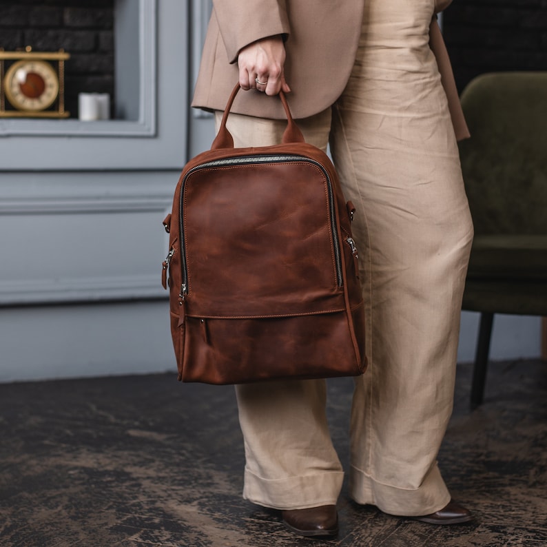 May include: A brown leather backpack with a zipper closure and adjustable straps. The backpack is being held by a person wearing a beige blazer and pants.