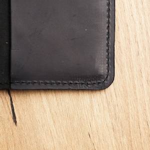 May include: Close-up of a black leather notebook with stitched edges. The notebook is lying on a light brown wooden surface.