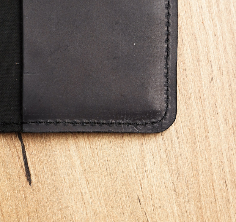 May include: Close-up of a black leather notebook with stitched edges. The notebook is lying on a light brown wooden surface.
