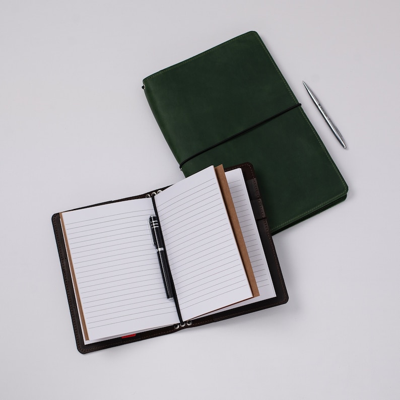 May include: Two leather journals, one open and one closed, with a pen. The open journal has lined pages and a black pen resting on it. The closed journal is dark green. A silver pen rests beside the green journal.
