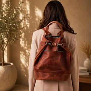 May include: A cognac-colored leather backpack featuring a top handle, adjustable shoulder straps, and a front exterior pocket with a horizontal zipper. The bag is secured with a metal clasp closure and includes silver-toned hardware accents.