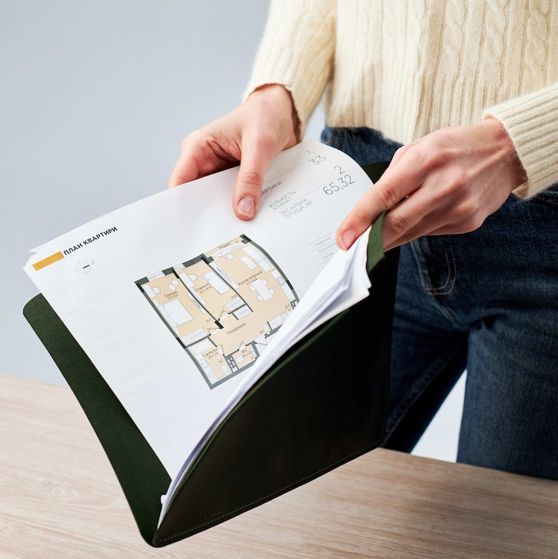 Op de afbeelding: Een persoon houdt een groene map vast met een wit vel papier erin. Het papier bevat een plattegrond met de tekst "План квартиры", wat in het Nederlands "Plattegrond van het appartement" betekent.