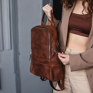 May include: A brown leather backpack with a zippered front pocket and adjustable straps. The backpack is being held by a person wearing a brown blazer and white pants.