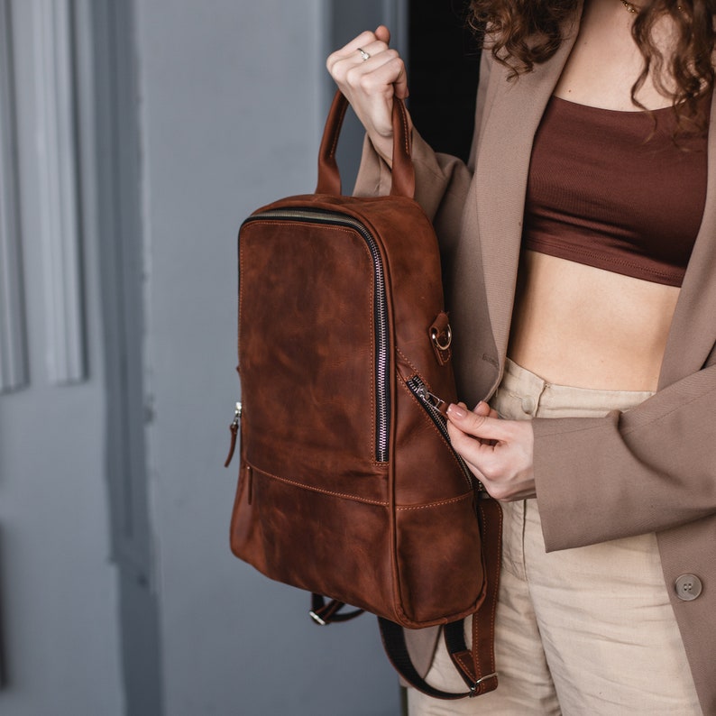 May include: A brown leather backpack with a zippered front pocket and adjustable straps. The backpack is being held by a person wearing a brown blazer and white pants.