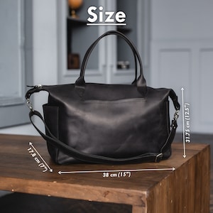 K&ouml;nnte beinhalten: Schwarze Ledertasche mit einem oberen Henkel und einem langen Schultergurt. Die Tasche misst 38 cm in der Breite, 31,75 cm in der H&ouml;he und 17,8 cm in der Tiefe.