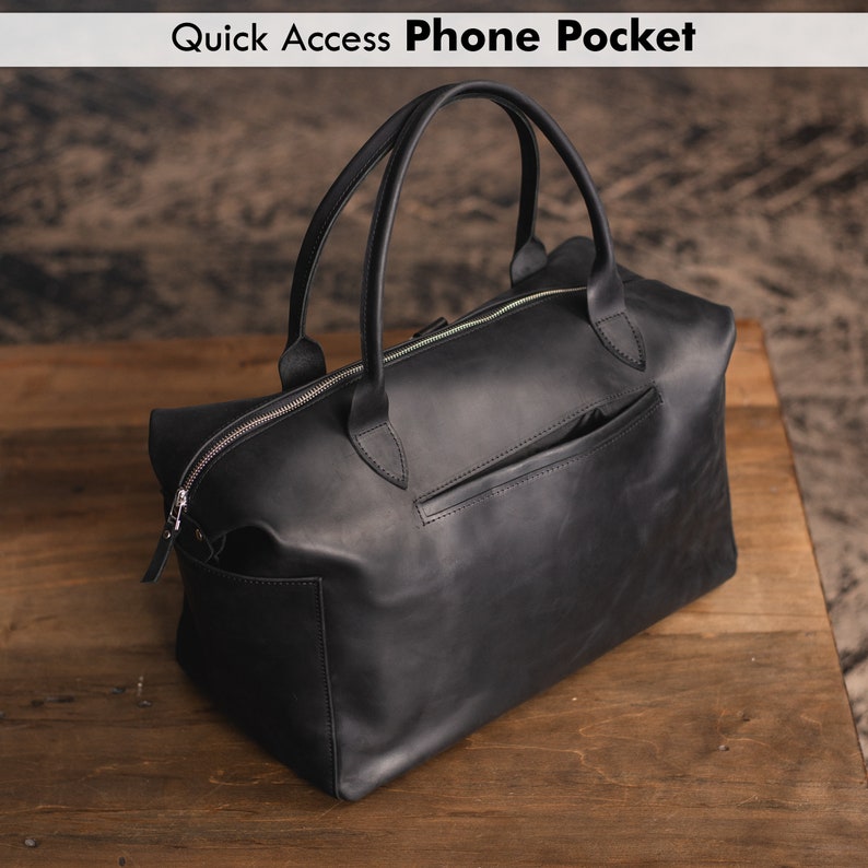 K&ouml;nnte beinhalten: Schwarze Leder-Reisetasche mit Rei&szlig;verschluss und einer kleinen Tasche auf der R&uuml;ckseite. Die Tasche hat zwei obere Griffe.