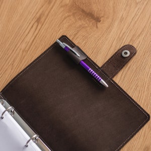 K&ouml;nnte beinhalten: Ein brauner Lederordner mit Stifthalter und einem lila-silbernen Stift. Der Ordner ist ge&ouml;ffnet und zeigt wei&szlig;es Papier im Inneren. Der Ordner hat einen Druckknopfverschluss und wei&szlig;e N&auml;hte.