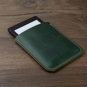 May include: A dark green leather e-reader sleeve with a contrasting light brown stitched border. The e-reader is partially visible, with a black frame and white screen. The sleeve is rectangular and sits on a wooden surface.