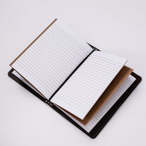 May include: Open brown leather journal with lined white pages. The journal has a black elastic band to keep it closed. The pages are lined and the journal is open, displaying the interior pages. The journal is on a white background.