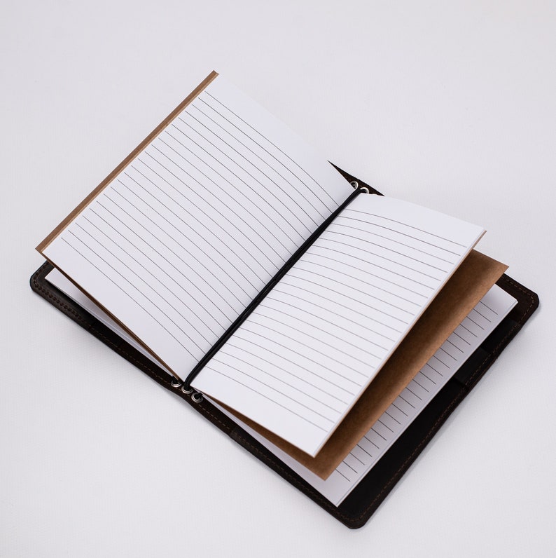May include: Open brown leather journal with lined white pages. The journal has a black elastic band to keep it closed. The pages are lined and the journal is open, displaying the interior pages. The journal is on a white background.