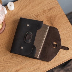 May include: A black leather portfolio with a brown flap and a silver buckle. The portfolio is open and a brown notebook is inside.