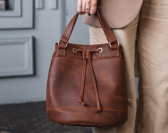 May include: A brown leather bucket bag with a drawstring closure and silver hardware. The bag has a top handle and is being held by a person.