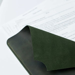 Op de afbeelding: Een donkergroene leren notitieboekomslag met een gestikte rand. De omslag is over een wit vel papier gevouwen.