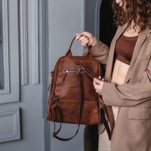 May include: A brown leather backpack with adjustable straps and a zippered pocket. The backpack is being held by a person.