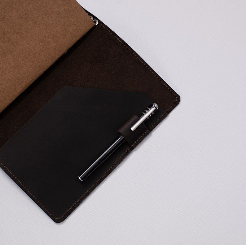 May include: A dark brown leather notebook cover with a black pocket and a silver pen. The notebook is open, revealing a tan paper insert. The cover has a stitched border and a pen holder.