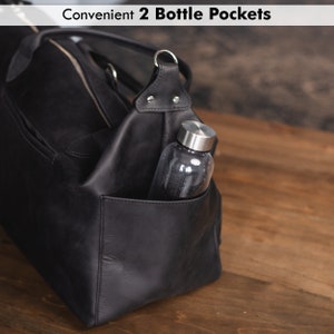 K&ouml;nnte beinhalten: Schwarze Leder-Reisetasche mit zwei Seitentaschen, eine mit einer Wasserflasche. Der Text "Convenient 2 Bottle Pockets" ist oben im Bild sichtbar.