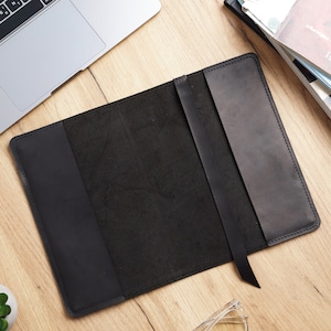 May include: Black leather notebook cover with a strap closure. The cover is lying flat on a wooden surface.