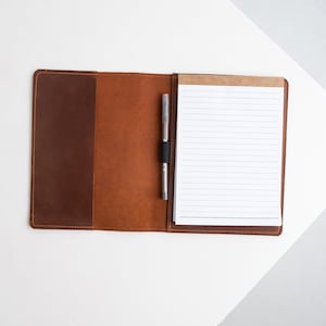 Pode incluir: Capa de bloco de notas de couro aberta em tons de castanho, com uma caneta presa por uma faixa elástica preta. O bloco de notas contém papel branco pautado. A capa está aberta, revelando o interior e o bloco de notas.