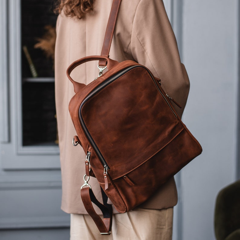May include: A brown leather backpack with silver zippers and buckles. The backpack has a flap with a zipper closure and a top handle.