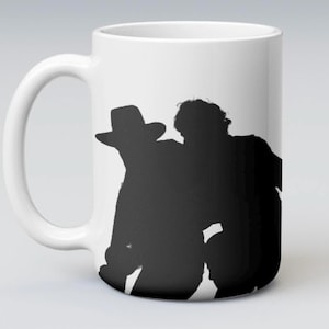 May include: A white ceramic mug featuring a black silhouette graphic of two figures, one wearing a wide-brimmed hat. The design is centered on the front of the mug, which has a curved handle on the left side and a smooth, cylindrical body.