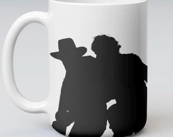 May include: A white ceramic mug featuring a black silhouette graphic of two figures, one wearing a wide-brimmed hat. The design is centered on the front of the mug, which has a curved handle on the left side and a smooth, cylindrical body.