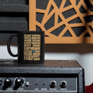 May include: A black ceramic mug featuring gold text that reads: HEART STOPPIN, PANTS DROPPIN, HARD ROCKIN, EARTH SHOCKIN, BOOTY SHAKIN, EARTHQUAKIN, LOVE MAKIN, TAKIN, HISTORY MAKIN, LEGENDARY E STREET BAND, with a small blue dot between MAKIN and TAKIN.
