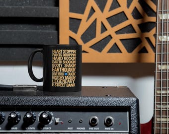 May include: A black ceramic mug featuring gold text that reads: HEART STOPPIN, PANTS DROPPIN, HARD ROCKIN, EARTH SHOCKIN, BOOTY SHAKIN, EARTHQUAKIN, LOVE MAKIN, TAKIN, HISTORY MAKIN, LEGENDARY E STREET BAND, with a small blue dot between MAKIN and TAKIN.