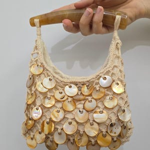 May include: A cream-coloured crochet handbag featuring a curved, translucent amber-toned handle. The body of the bag is adorned with numerous circular, iridescent shell discs in shades of brown and gold, attached to the open-weave fabric.