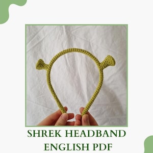 May include: A green crocheted headband with two ears, resembling Shrek's ears. The headband is held in two hands. The text "SHREK HEADBAND ENGLISH PDF PATTERN" is below the headband.