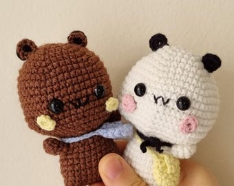 May include: Two handmade crochet amigurumi dolls. One is brown with a blue scarf and yellow accents, the other is white with black panda-like features and a yellow bag. Both have black eyes and pink cheeks.