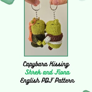 May include: Two handmade crochet keychains depicting Shrek and Fiona kissing. The Shrek keychain is green with a brown vest, and the Fiona keychain has orange hair. The keychains are held up by a person's fingers. The text "Capybara Kissing Shrek and Fiona English PDF Pattern" is below.