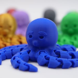 May include: A blue 3D printed octopus with a cartoon-like face. The octopus has eight tentacles and is sitting on a white surface.