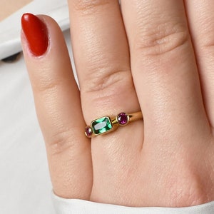 Peut inclure: Une bague en or avec une émeraude verte et deux petits rubis rouges. La bague est portée sur un doigt avec un vernis à ongles rouge.