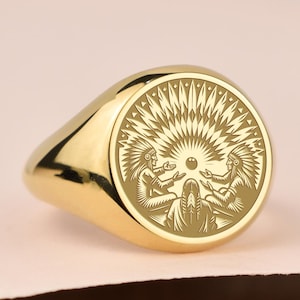 May include: A gold signet ring with an engraved design. The ring features a circular face with a detailed illustration of figures and sunburst elements. The ring is displayed on a light pink surface.