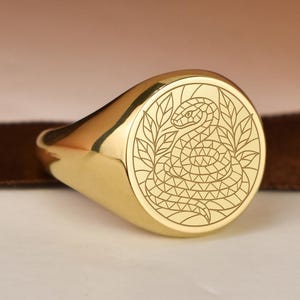 Może przedstawiać: Złoty sygnet z szczegółowym wzorem węża. Pierścień ma polerowane wykończenie i okrągłą powierzchnię z emblematem węża. Wąż otoczony jest wzorem wieńca laurowego. Pierścień jest umieszczony na neutralnym tle.