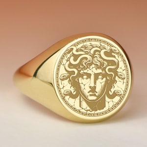 Può includere: Anello con sigillo dorato con un'incisione dettagliata del volto di Medusa. L'anello ha una finitura liscia e lucida e una forma ovale arrotondata. L'incisione di Medusa è centrata sulla faccia dell'anello.