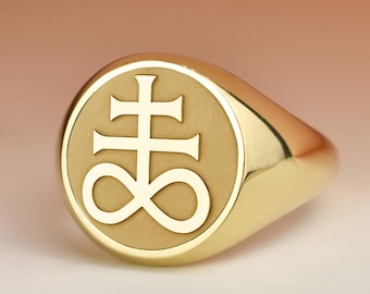 May include: A gold-colored signet ring with a circular face. The face features a stylized cross above an infinity symbol. The ring is smooth and polished, reflecting light. The ring is a symbol of the occult.