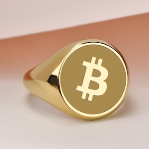 May include: A gold-colored signet ring featuring the Bitcoin symbol. The ring has a rounded face with a light brown background and a white Bitcoin logo. The band is smooth and polished.