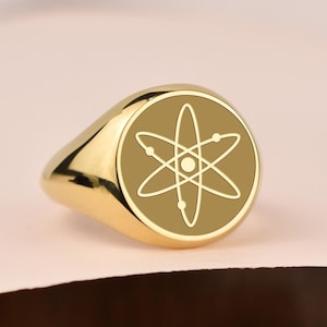 Op de afbeelding: Een goudkleurige zegelring met een cirkelvormige voorkant met een wit atoomteken. De ring heeft een gepolijste afwerking en staat tegen een zachte, neutrale achtergrond. Het ontwerp van de ring is eenvoudig en elegant.