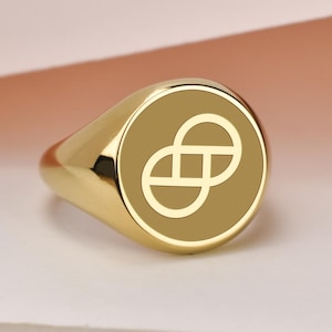 May include: A gold signet ring with a circular face. The face features a light-colored, stylized infinity symbol on a darker gold background. The ring is highly polished and reflects the light.