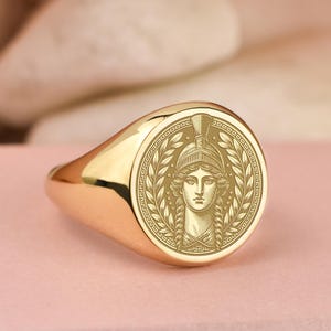 Puede incluir: Un anillo de sello dorado con un diseño grabado detallado. El anillo presenta una cara redonda con la representación de una mujer con casco, rodeada por una corona de laurel. El anillo está sobre una superficie rosa.