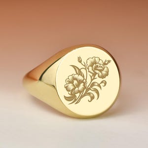 Può includere: Un anello con sigillo dorato con un motivo floreale inciso sulla superficie ovale. L'anello ha una finitura lucida e una fascia arrotondata. Il motivo floreale presenta due fiori con foglie e steli.