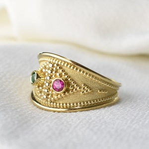 May include: A gold ring with a textured band and two gemstones. The ring features a small green gemstone and a larger pink gemstone. The ring has a detailed design with small beaded accents.