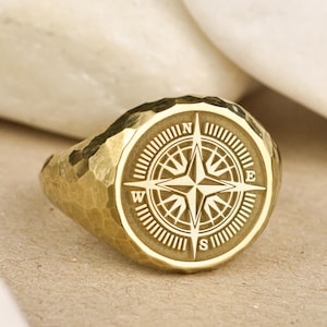 May include: A gold signet ring with a compass design engraved on the top. The compass features a star in the center with the letters N, S, E, and W around the edge.