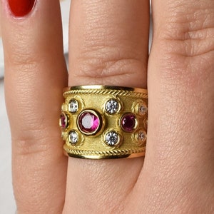 May include: A wide gold ring with a textured surface, adorned with alternating round red and clear gemstones. The ring is on a hand with red nail polish, set against a white shirt.