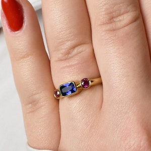 Peut inclure: Une bague en or avec une pierre rectangulaire bleue et deux petites pierres roses. La bague est portée sur un doigt avec un vernis à ongles rouge.