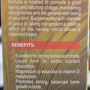 May include: A product description on a brown and gold box for a nutritional supplement for birds. The text highlights benefits like strong bones, better nutrient absorption, and shiny plumage. The recommended dose is also included.